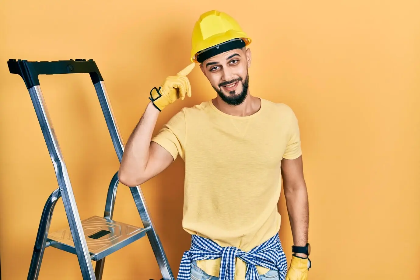 A handsome bearded man beside construction stairs, wearing a hardhat, smiling, and pointing to his head with one finger, suggesting a great idea or good memory.